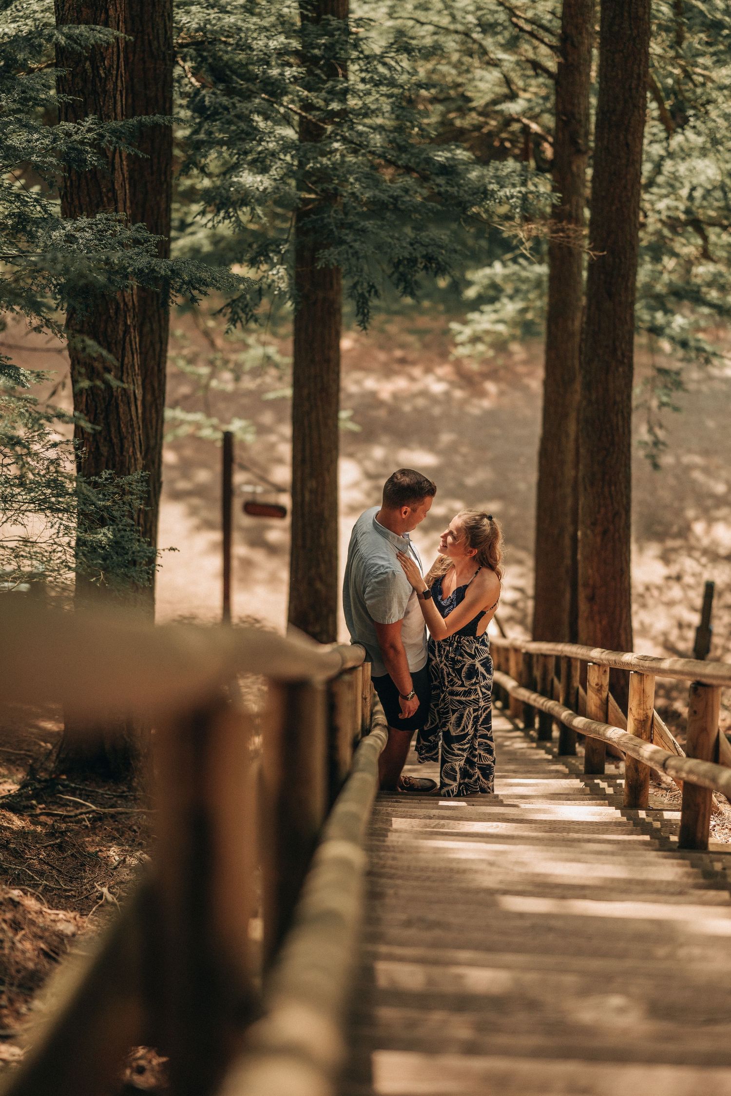 A couple shares an intimate moment on a wooden boardwalk surrounded by towering trees.