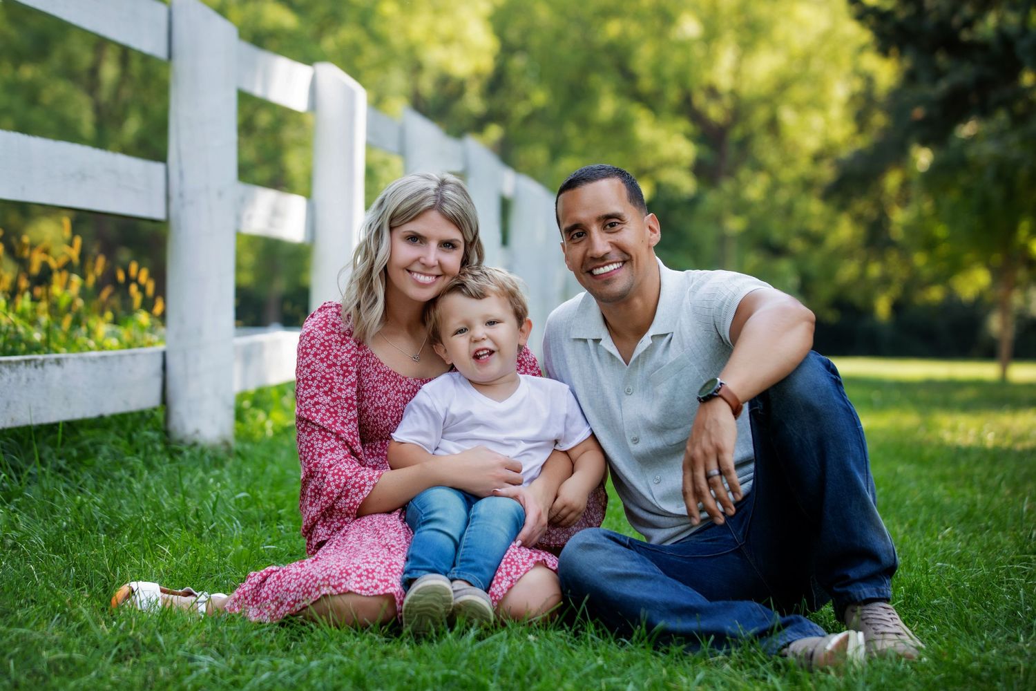 Family sitting together on grass by white fence in sunny outdoor setting.