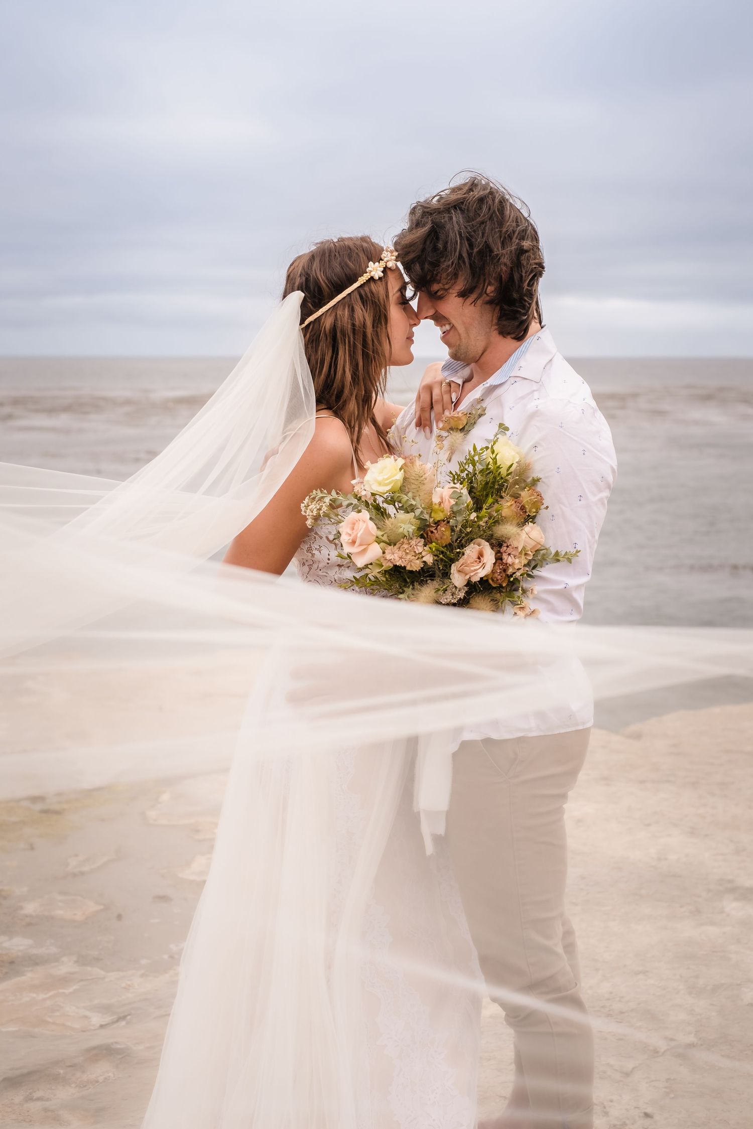 A romantic beach wedding scene with a flowing white veil catching the wind as a couple embraces on an overcast day. Santa Cruz CA