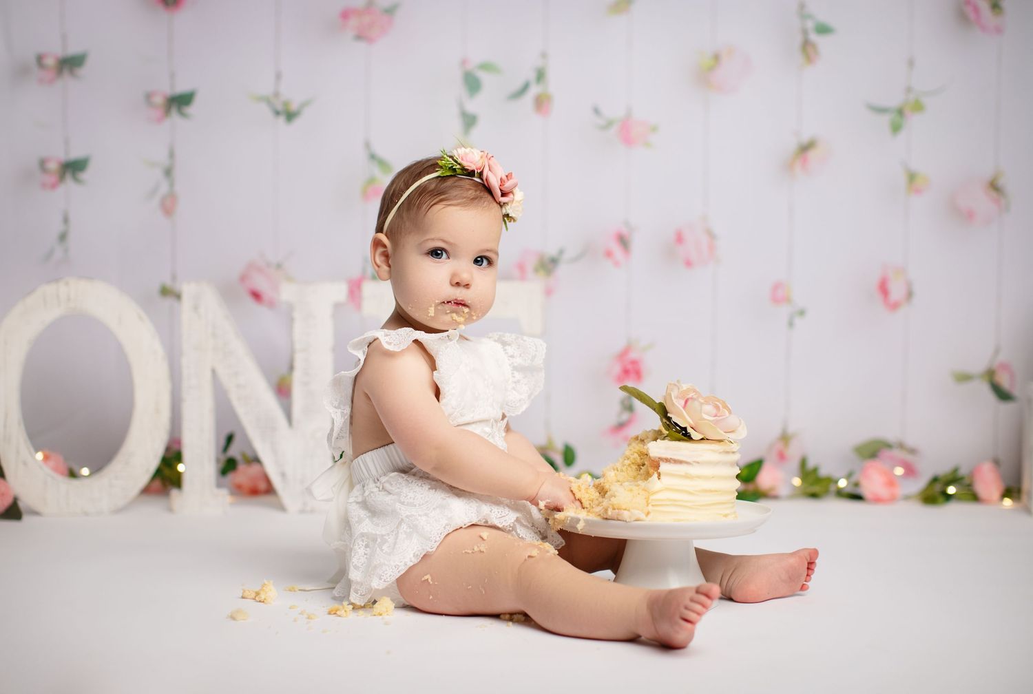 Baby in white dress sitting next to 'ONE' letters and cake for first birthday photoshoot.