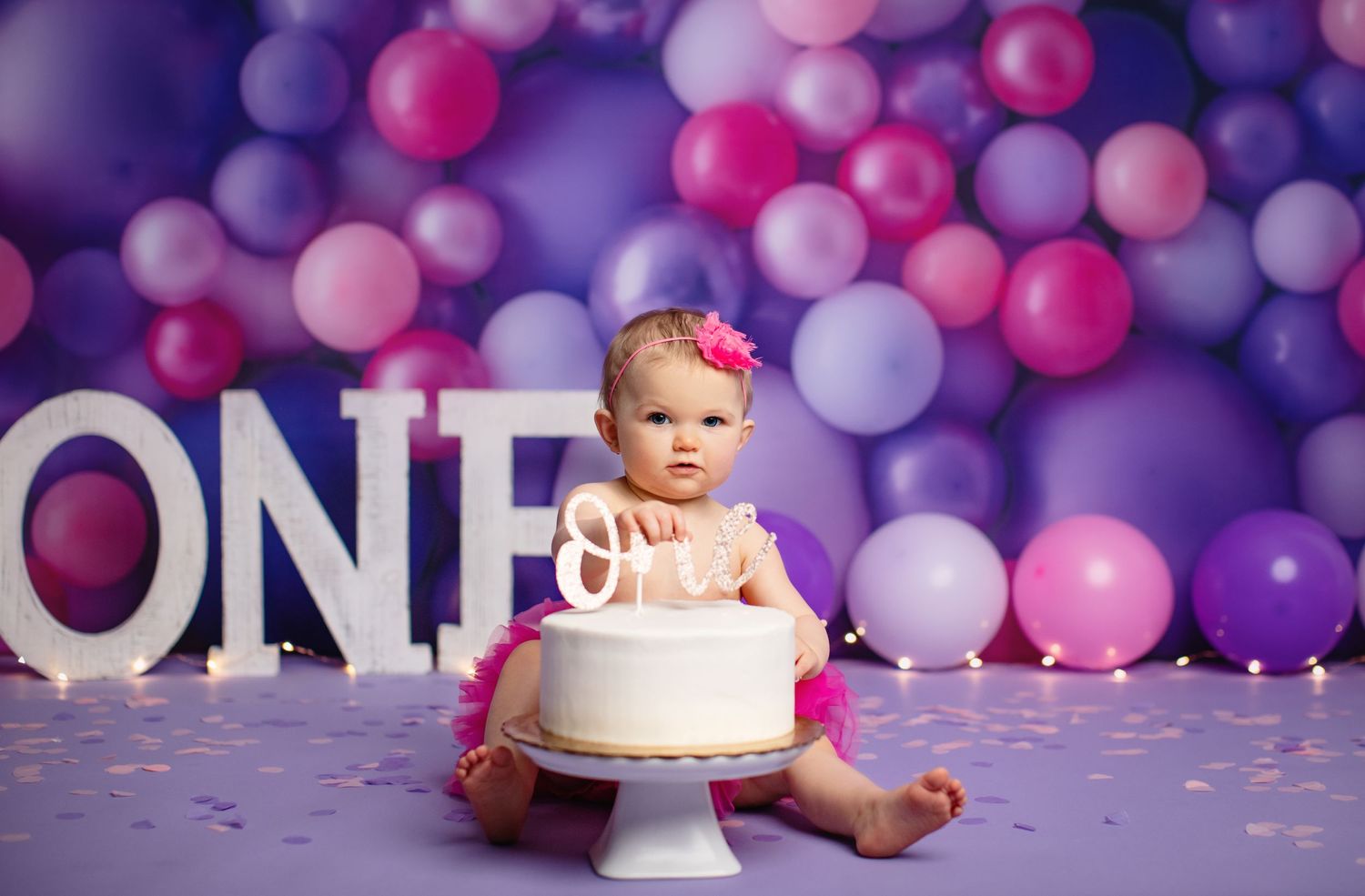 Baby with birthday cake against purple and pink balloon backdrop with 'ONE' letters.
