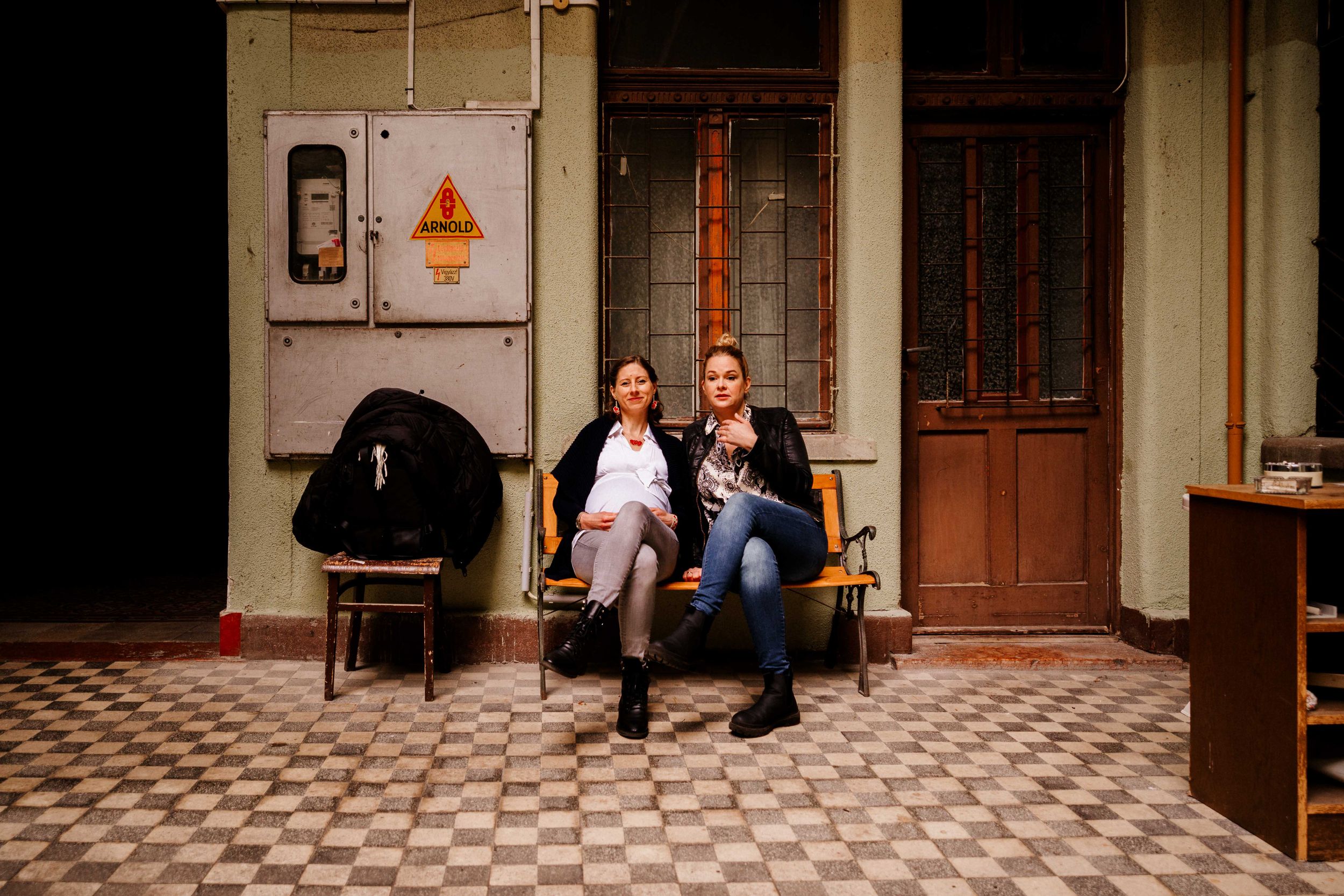 Two people sitting and chatting on a bench outside a building with patterned floor tiles.