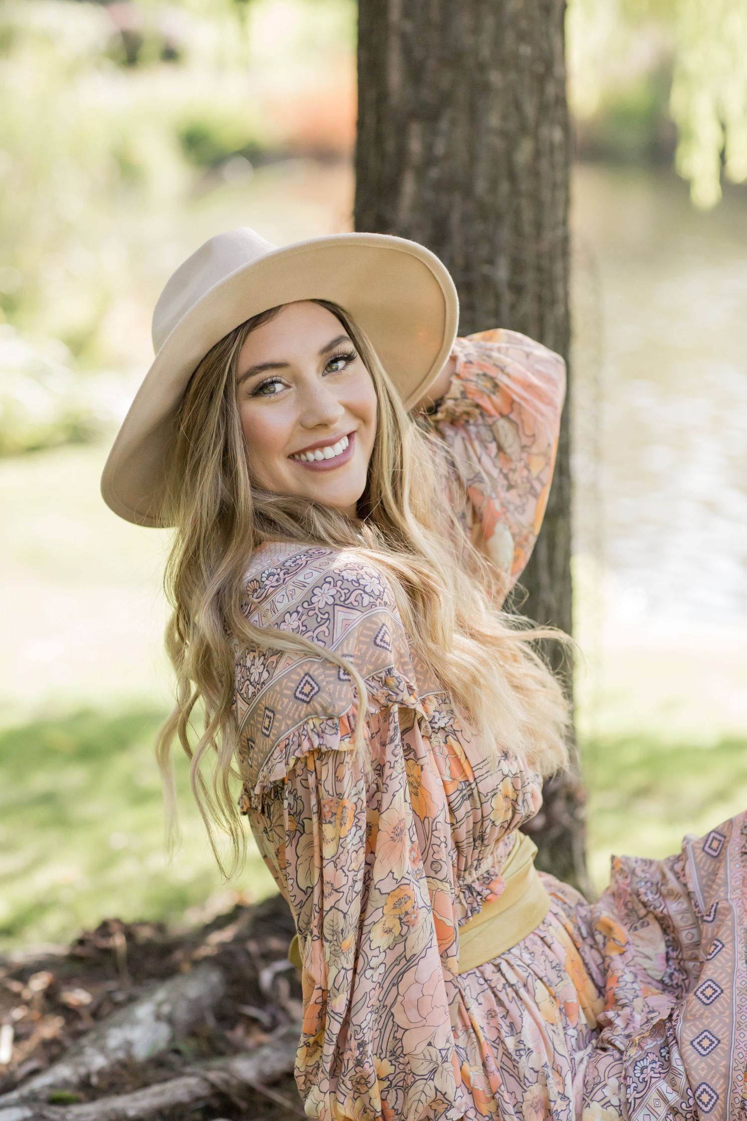 Smiling blonde woman in bohemian floral dress and tan hat leaning against a tree near a serene lake.