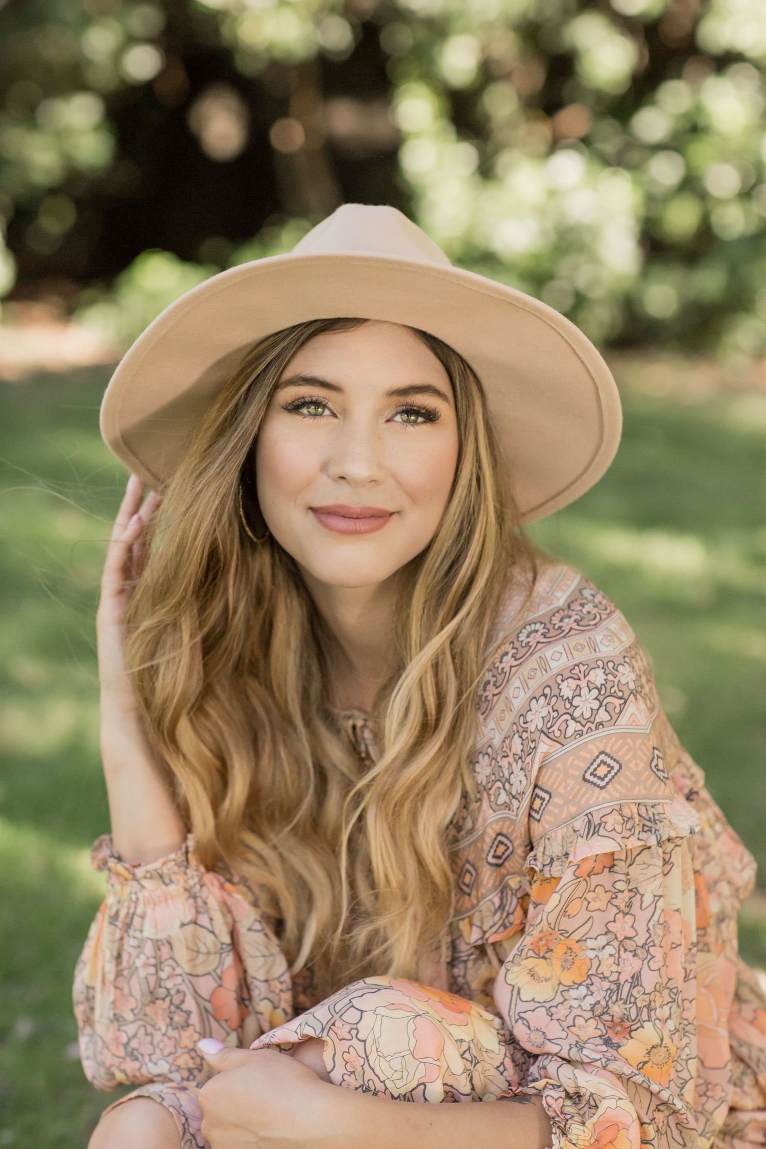Young blonde woman in floral boho dress and tan wide-brim hat sitting in green park.
