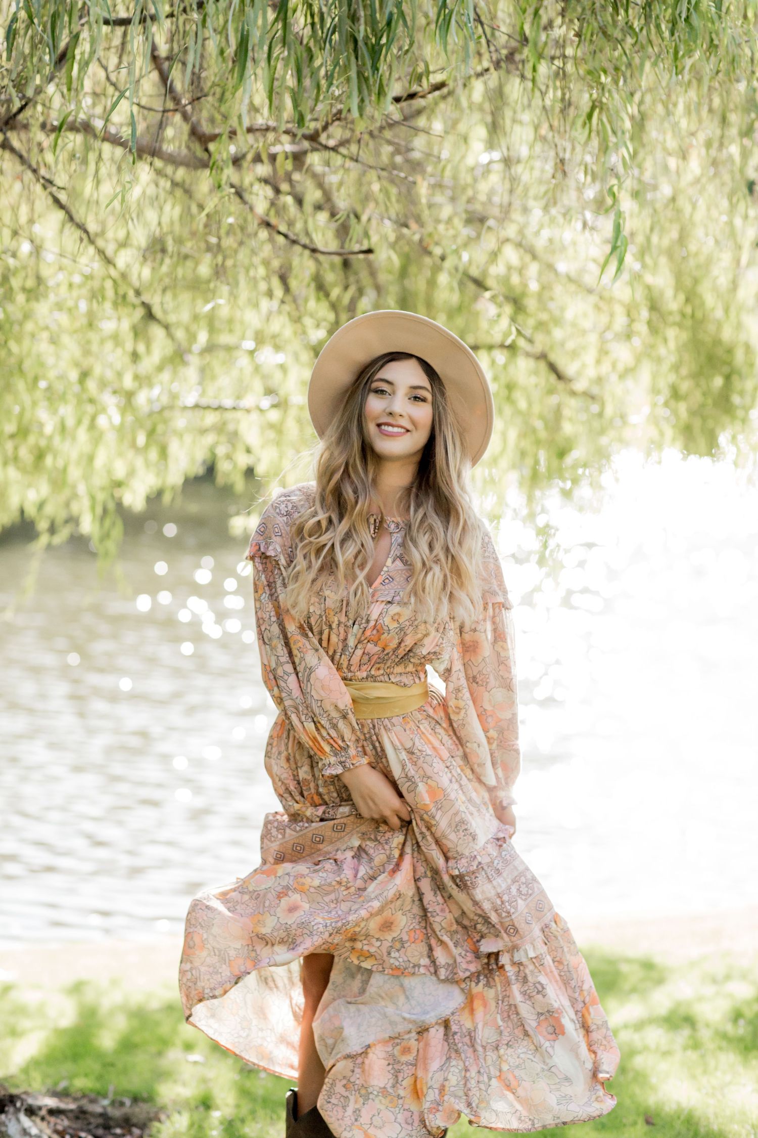 Young woman in floral maxi dress and tan wide-brim hat smiling by a sunlit lake under willow trees.