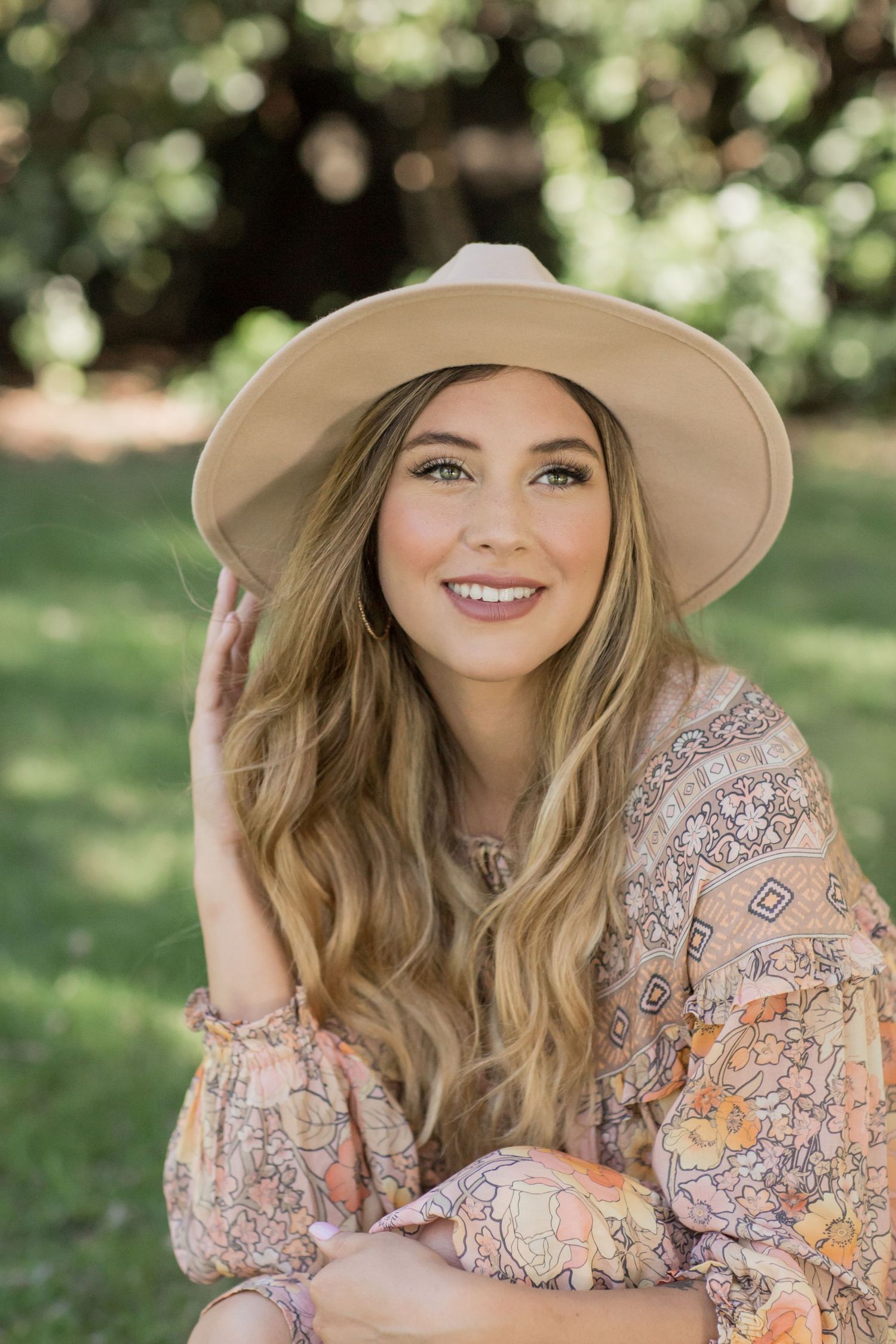 Smiling blonde woman in floral boho top wearing a wide-brim tan hat while sitting outdoors in a park.
