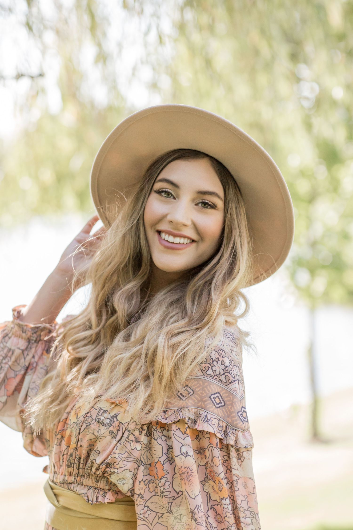 Cheerful blonde woman in floral top and tan wide-brim hat posing outdoors near a sunlit willow tree.