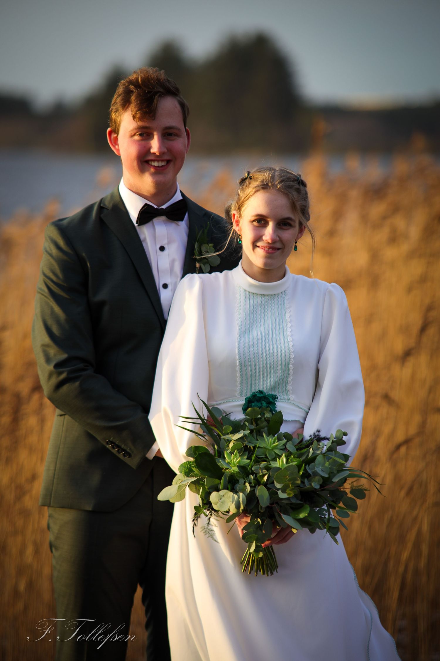 Smiling bride and groom posing together near golden reeds holding a green bouquet at sunset.