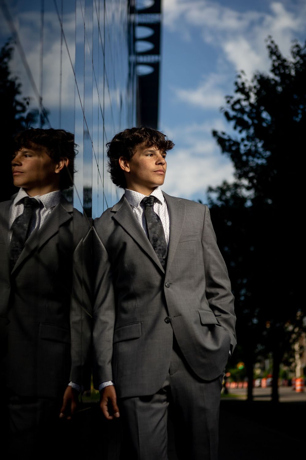 A young man in a gray suit poses against a reflective glass surface.
