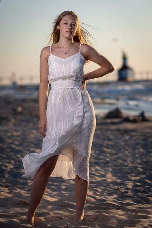Person in white sundress poses on beach at sunset with wind-blown fabric.