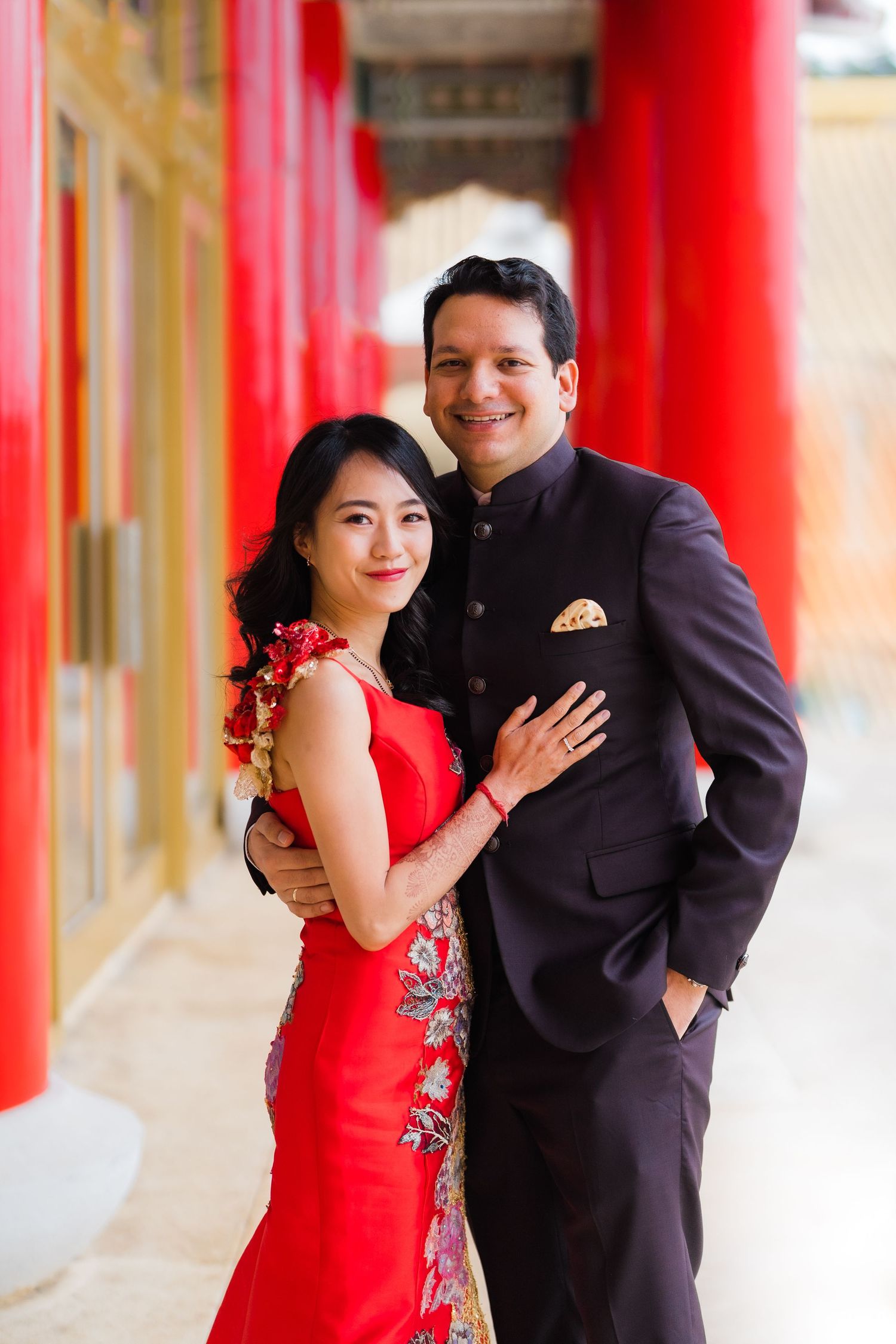 A couple poses in formal attire against red pillars, wearing a red dress and black suit.