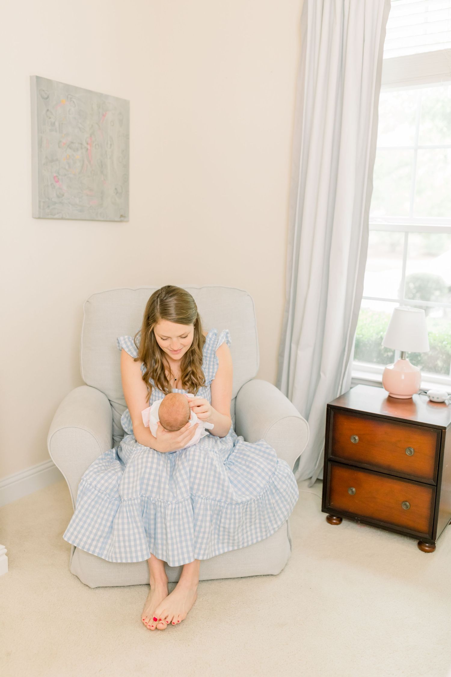 A woman in a blue gingham dress sits in a light gray armchair in a minimalist cream-colored room.
