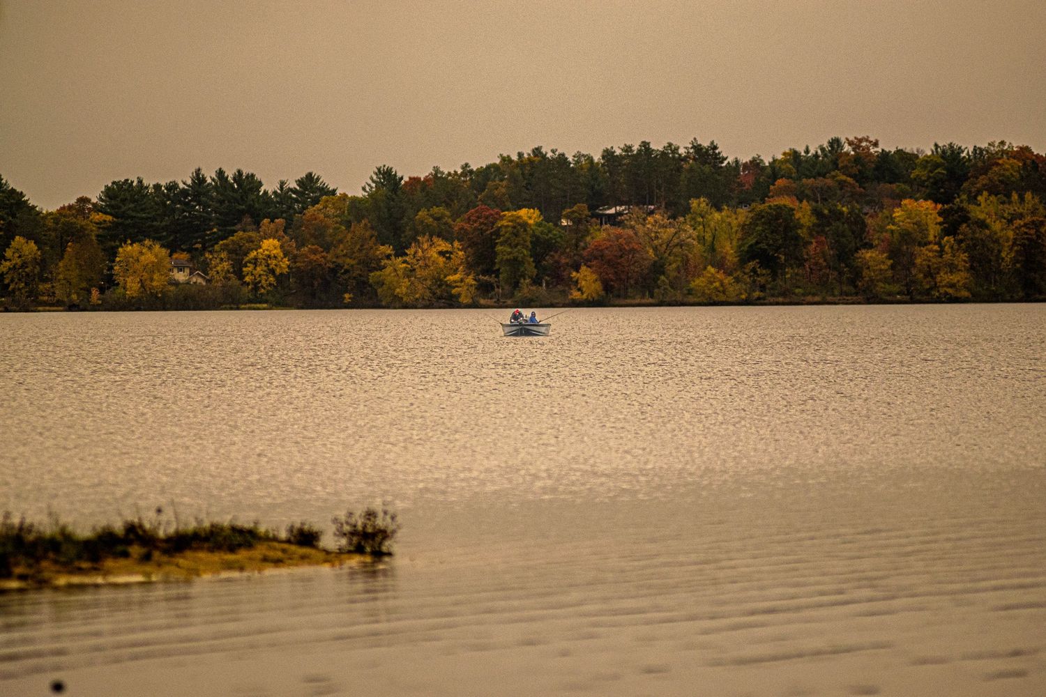 A small boat floats on a tranquil lake surrounded by autumn trees with golden foliage at sunset.