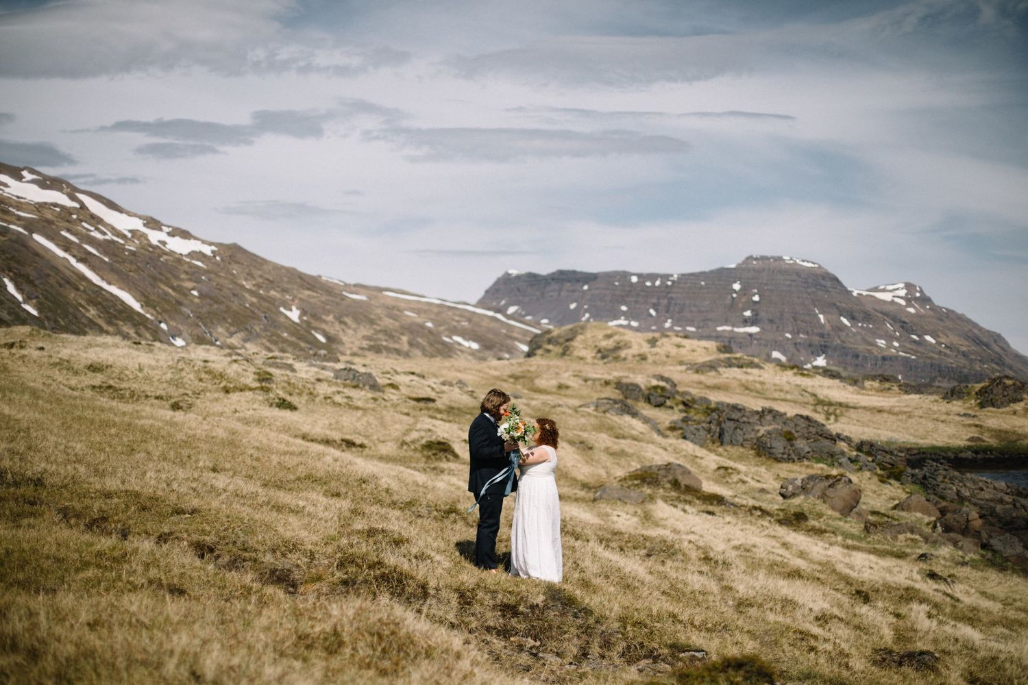 Couple in wedding attire embracing on mountainside with cloudy sky.