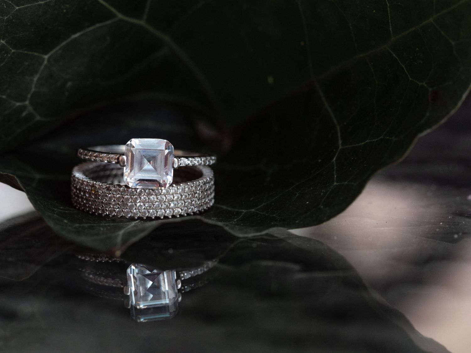 Elegant emerald-cut diamond engagement ring with pave band reflecting on dark glossy surface with green leaf backdrop.