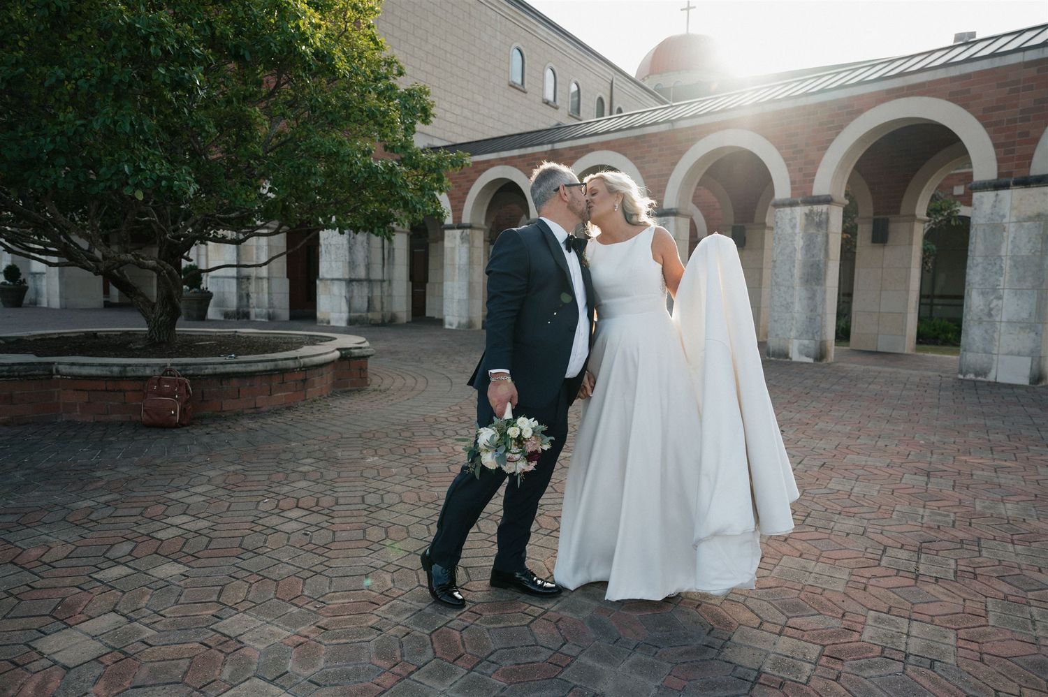 A couple in wedding attire share a romantic moment under stone arches in a courtyard at sunset.