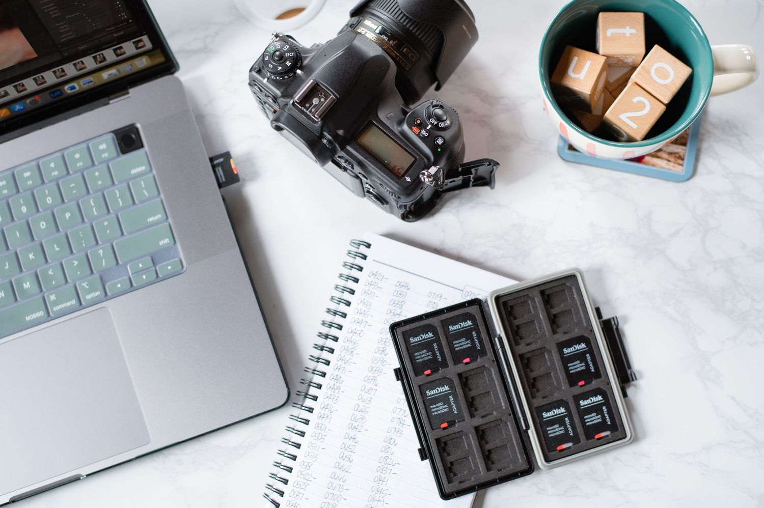Flatlay of photography equipment with laptop, camera, memory card case and snacks.