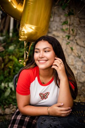 A person wearing a red and white t-shirt with a butterfly design smiles while sitting outdoors with golden balloons.