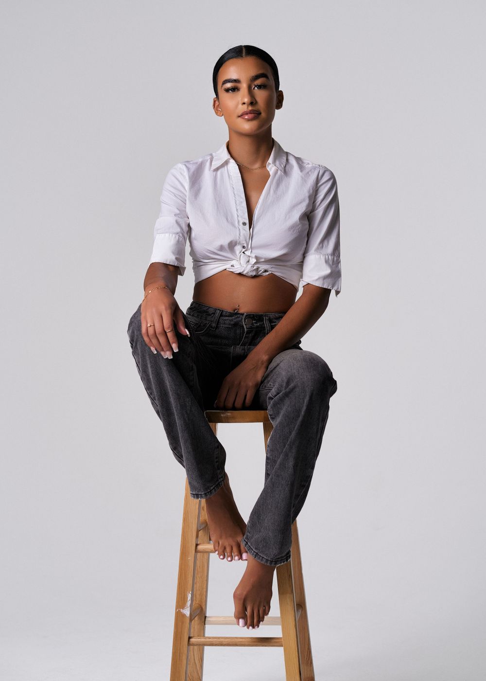 Model in cropped white shirt and jeans poses casually on wooden stool.
