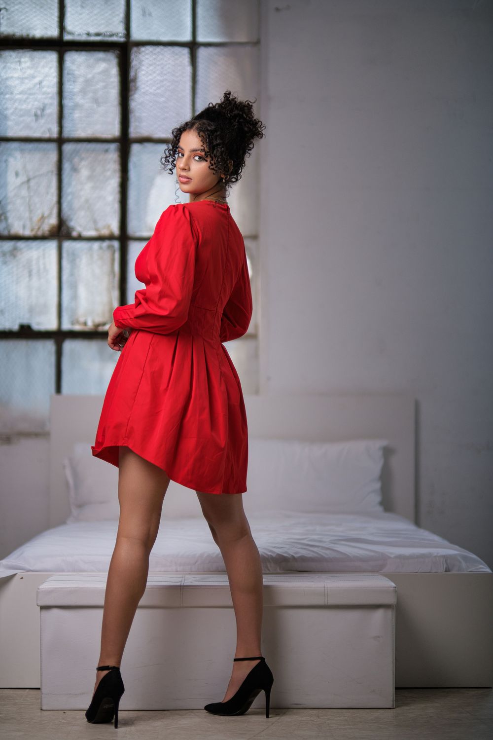 Model strikes dynamic pose in short red dress and heels against white studio backdrop.