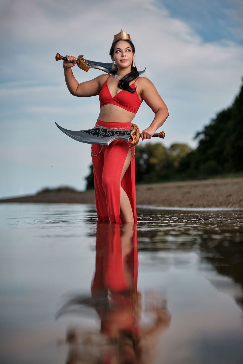 Warrior poses with sword in flowing red dress at water's edge with dramatic reflection.