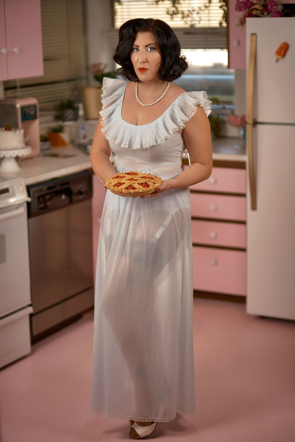 A retro kitchen scene with pink appliances featuring a white vintage nightgown and pearl necklace.