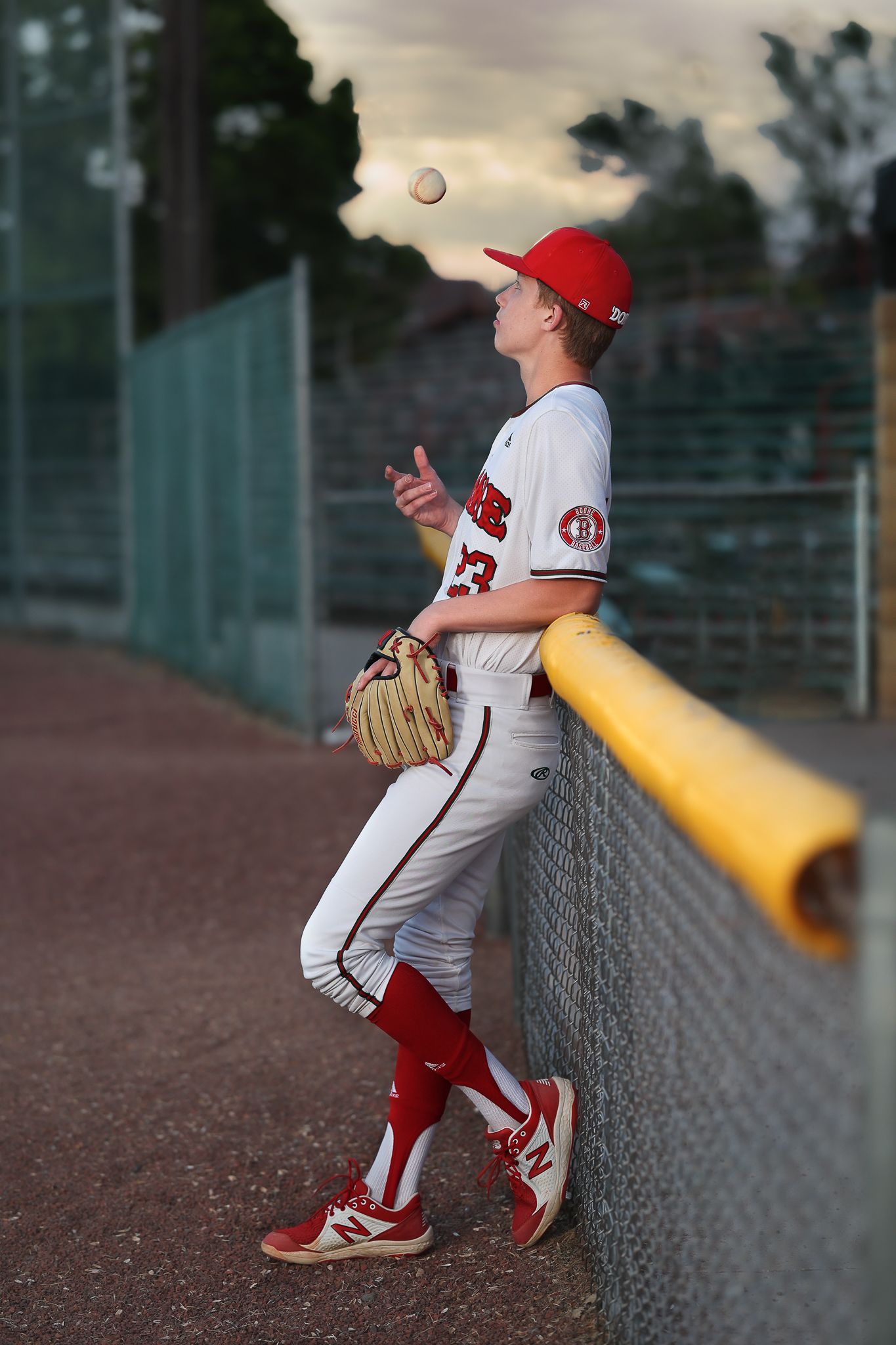 Baseball player in uniform leaning against fence while tossing ball at sunset.
