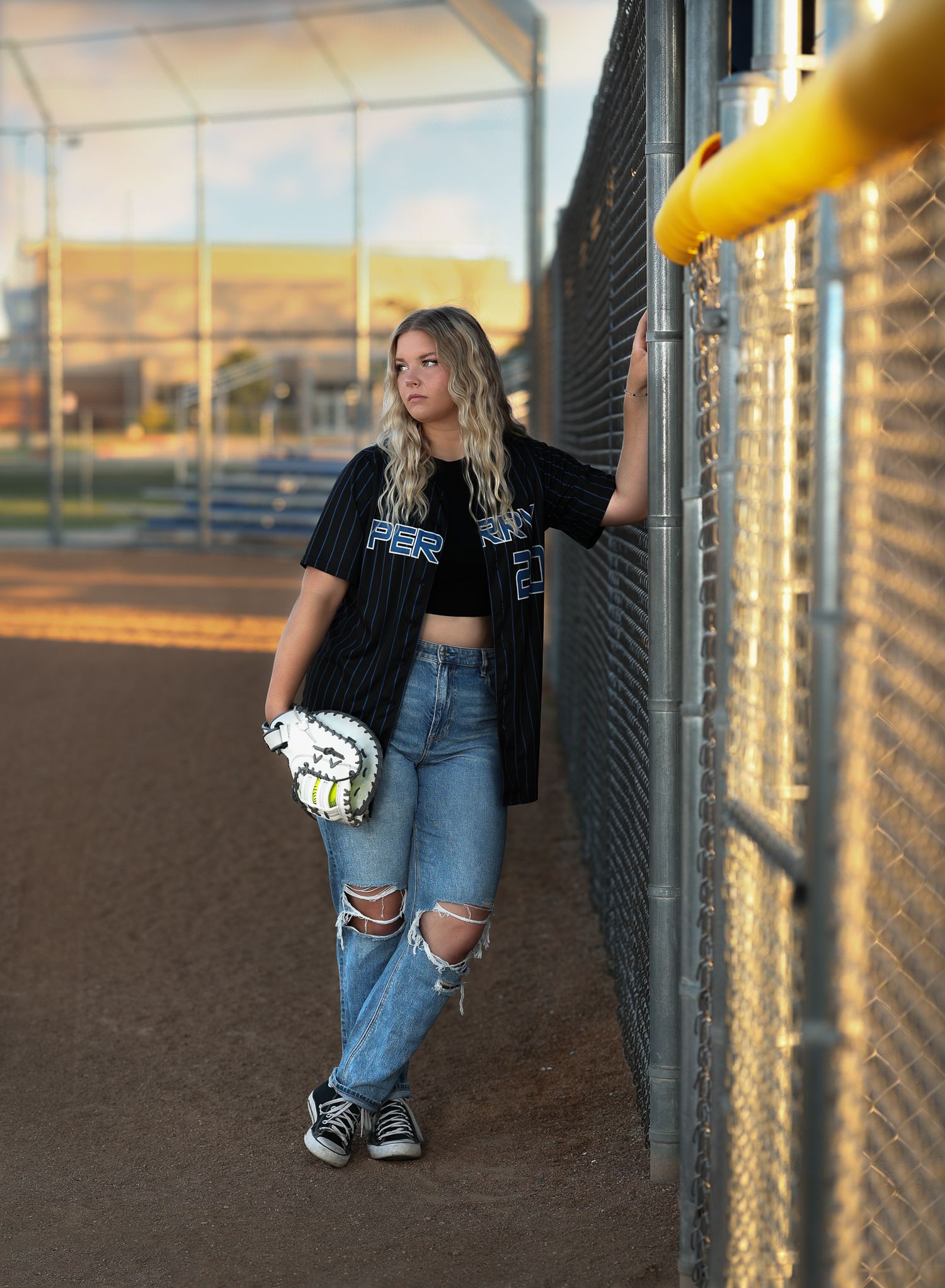 Softball player in ripped jeans and team jersey poses against fence during golden hour.