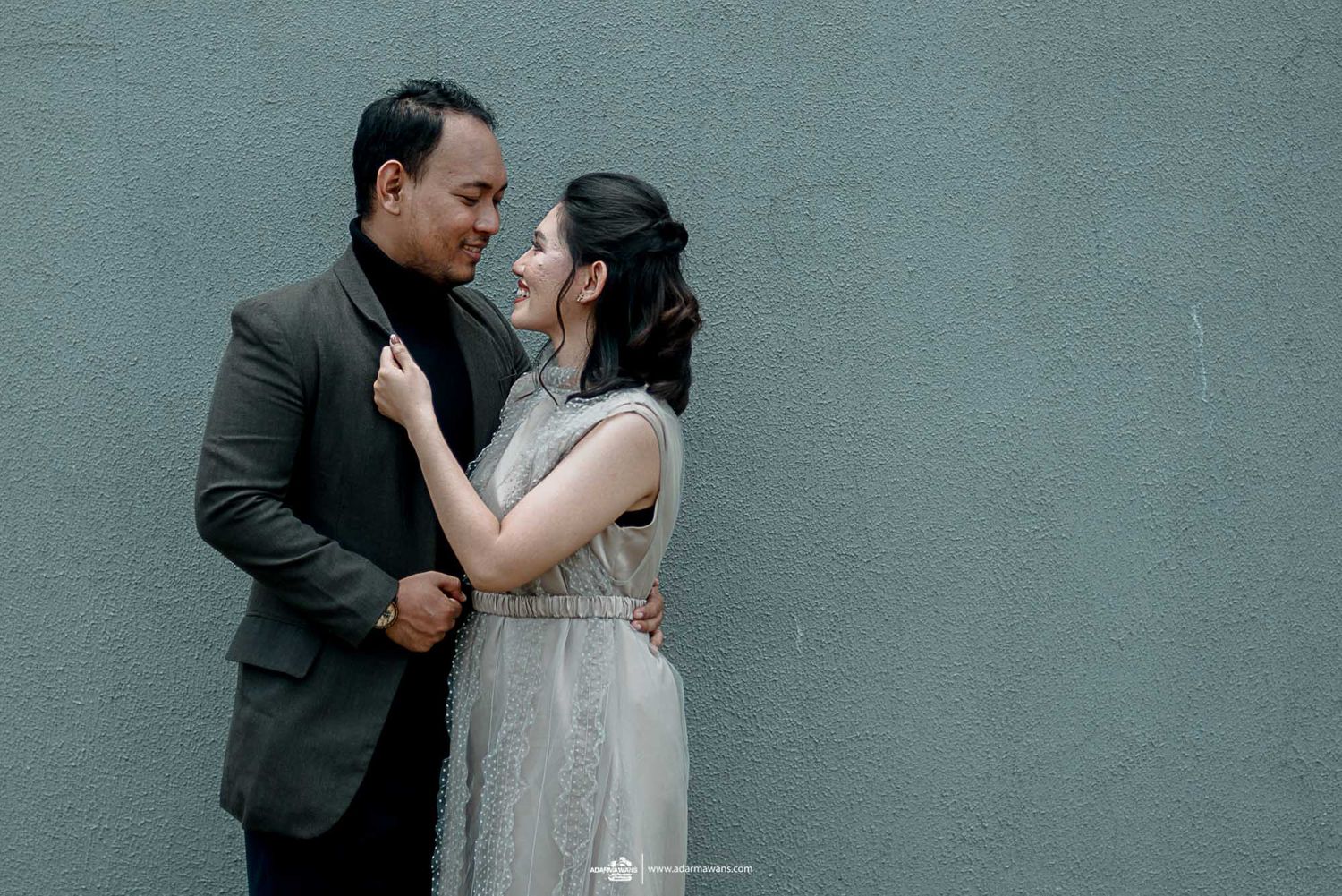A romantic couple embraces against a grey wall in formal attire.