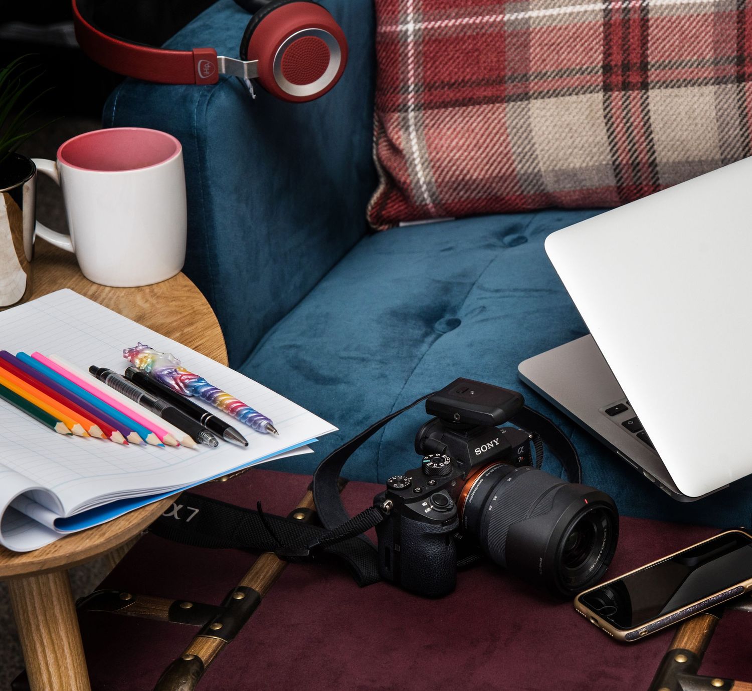 Camera equipment and supplies arranged on a plaid blanket includes lenses, writing tools, and a coffee cup.