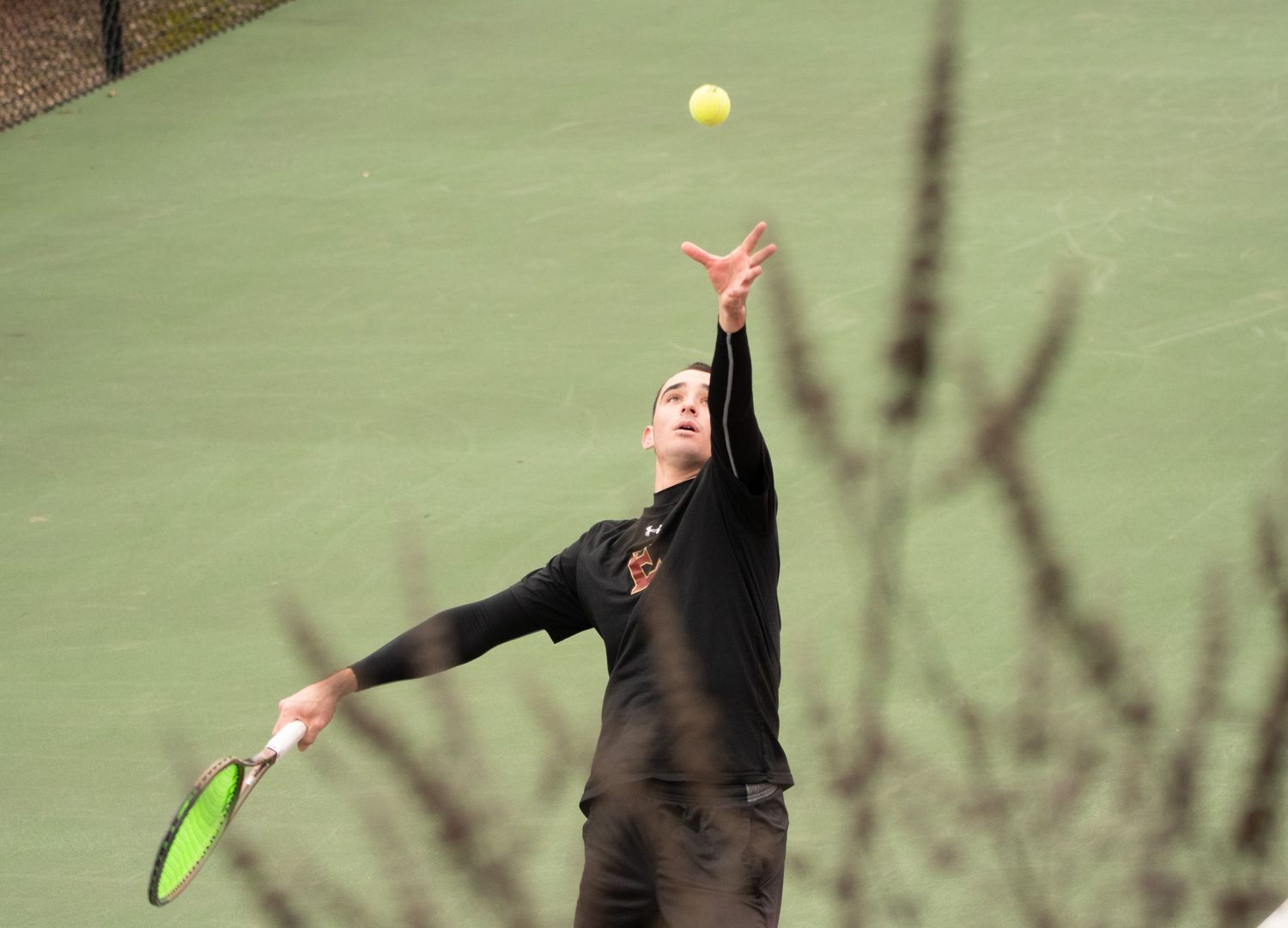 Tennis player reaches up to serve the ball on a green court.