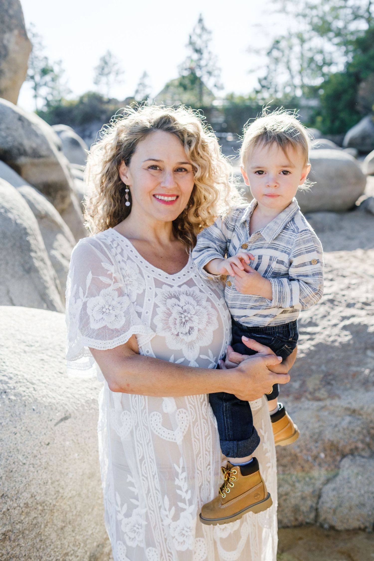 A pregnant woman stands outdoors holding a toddler wearing a striped sweater and boots, with rock formations in the background.