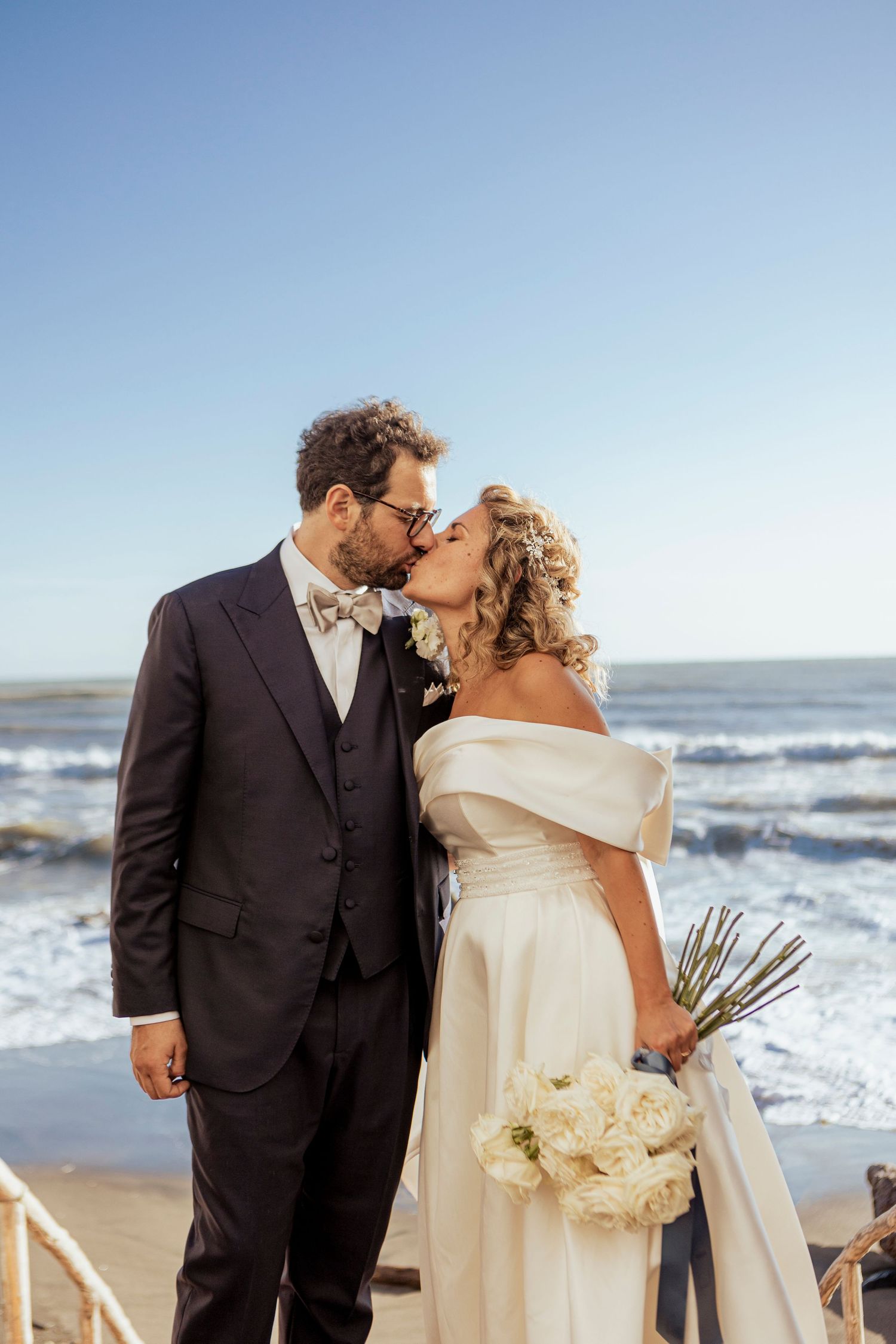 Romantic seaside wedding portrait with couple sharing a kiss against ocean backdrop.