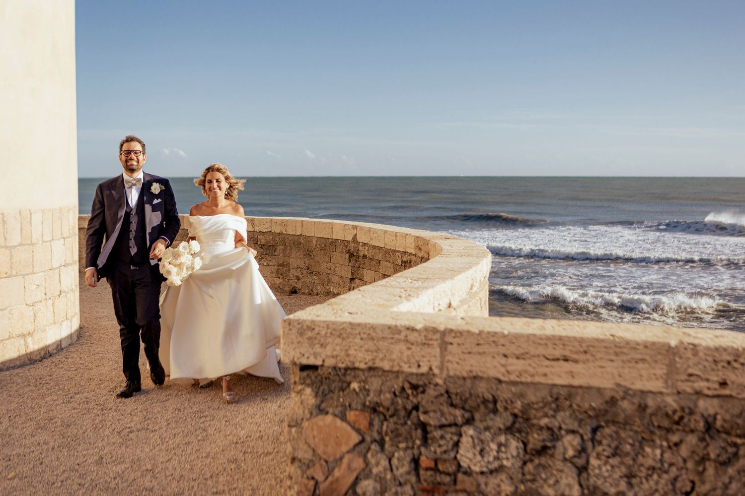Bride and groom walking along stone oceanfront path during destination wedding.