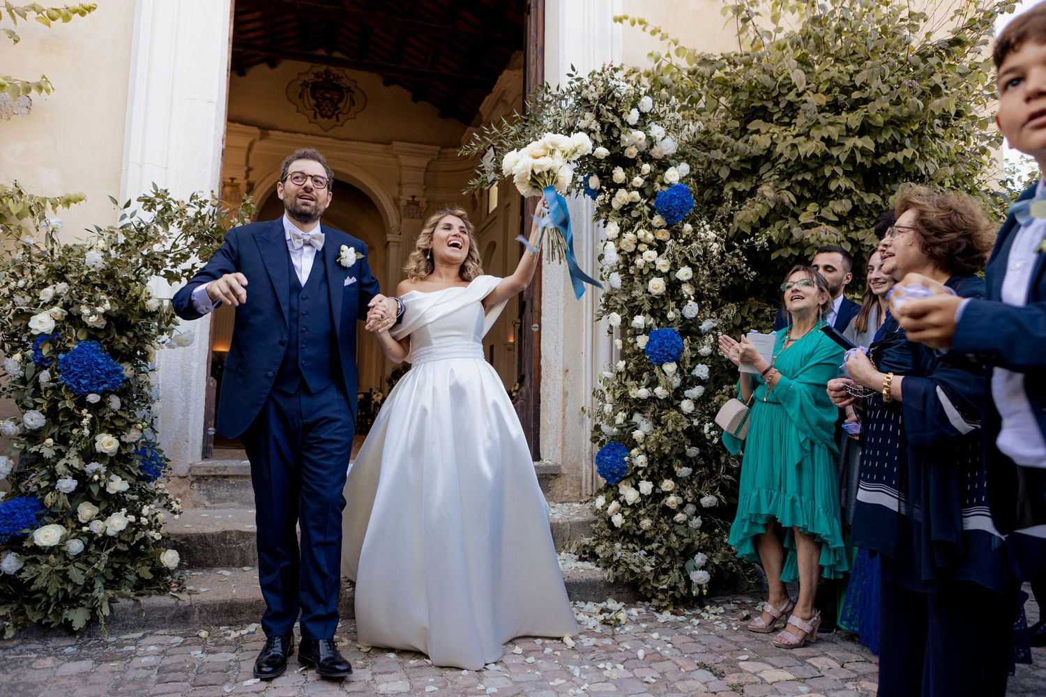 Wedding couple exits church through floral arch as guests shower them with confetti.