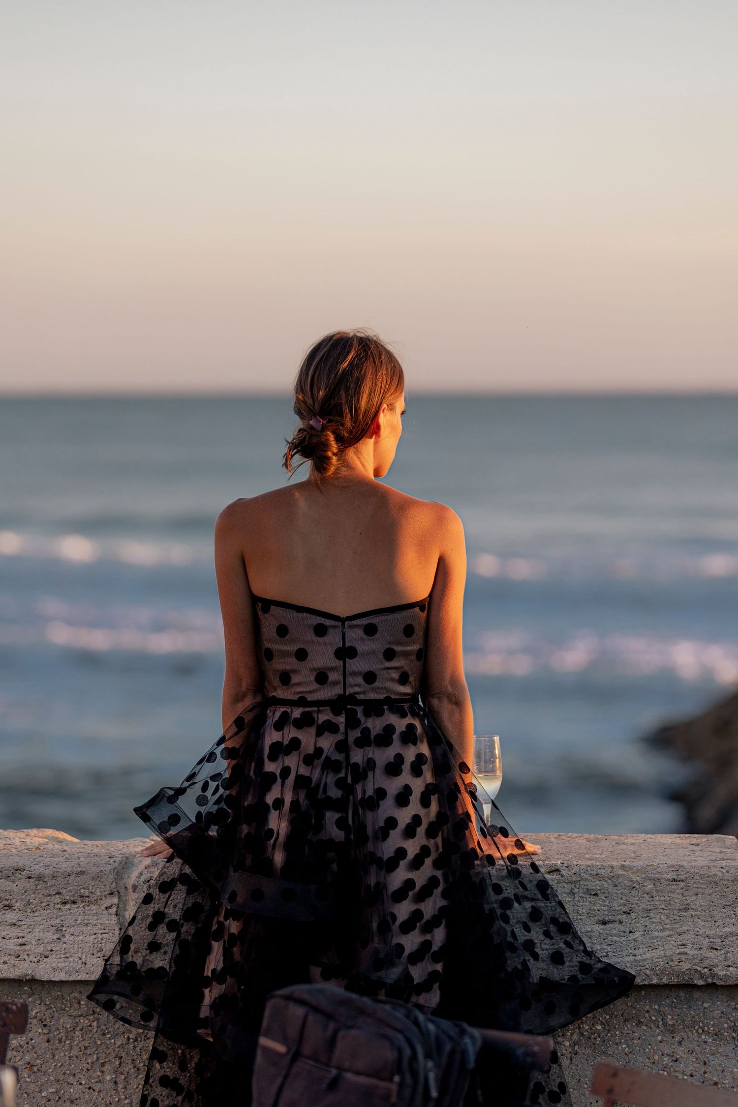 Woman in polka dot dress sits watching the sunset over the ocean.