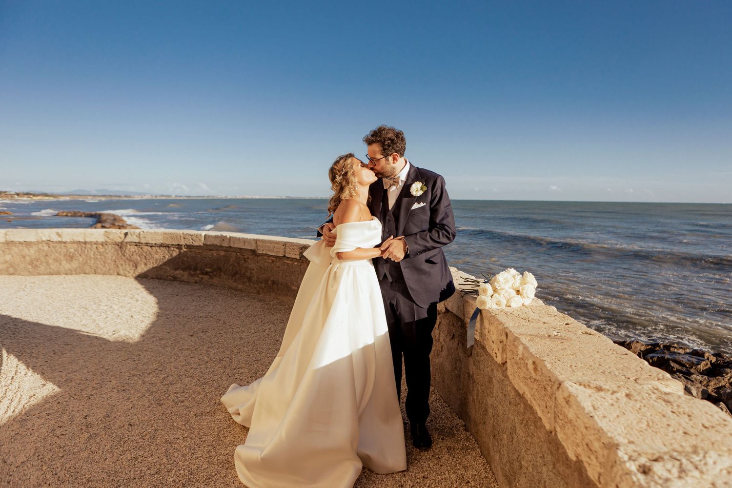Couple in wedding attire embracing on stone balcony overlooking ocean view.