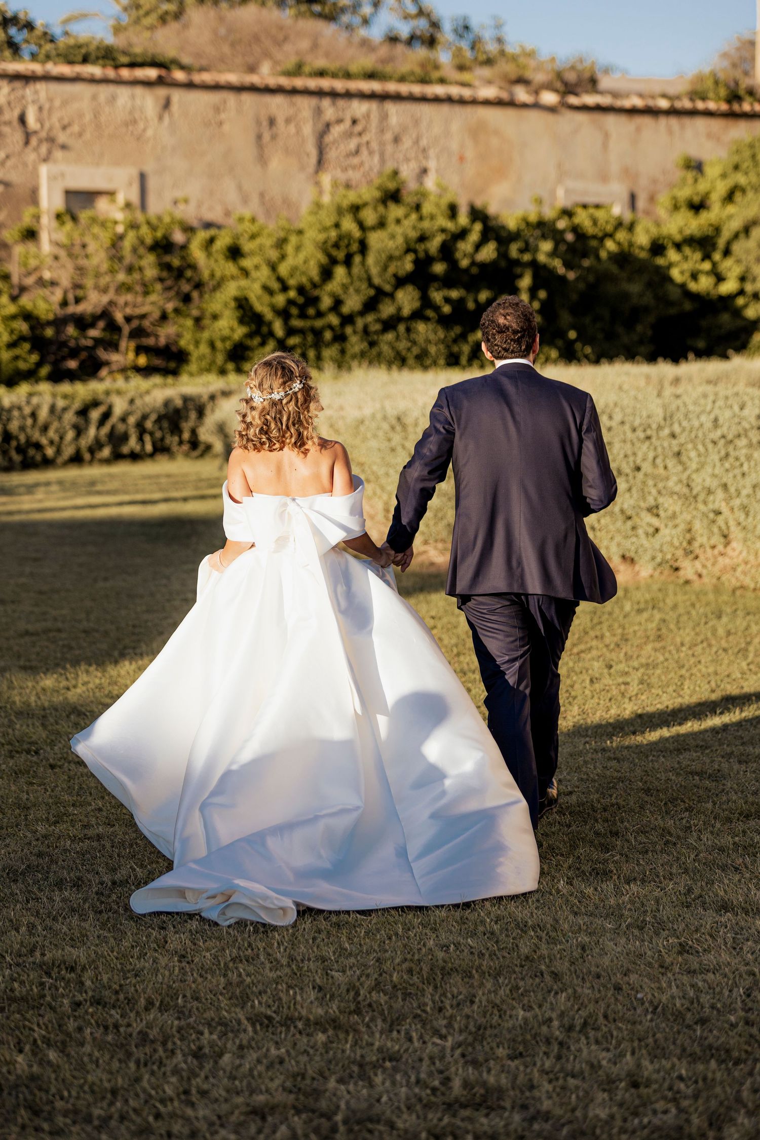 Couple in wedding attire walking together across a manicured lawn at sunset.