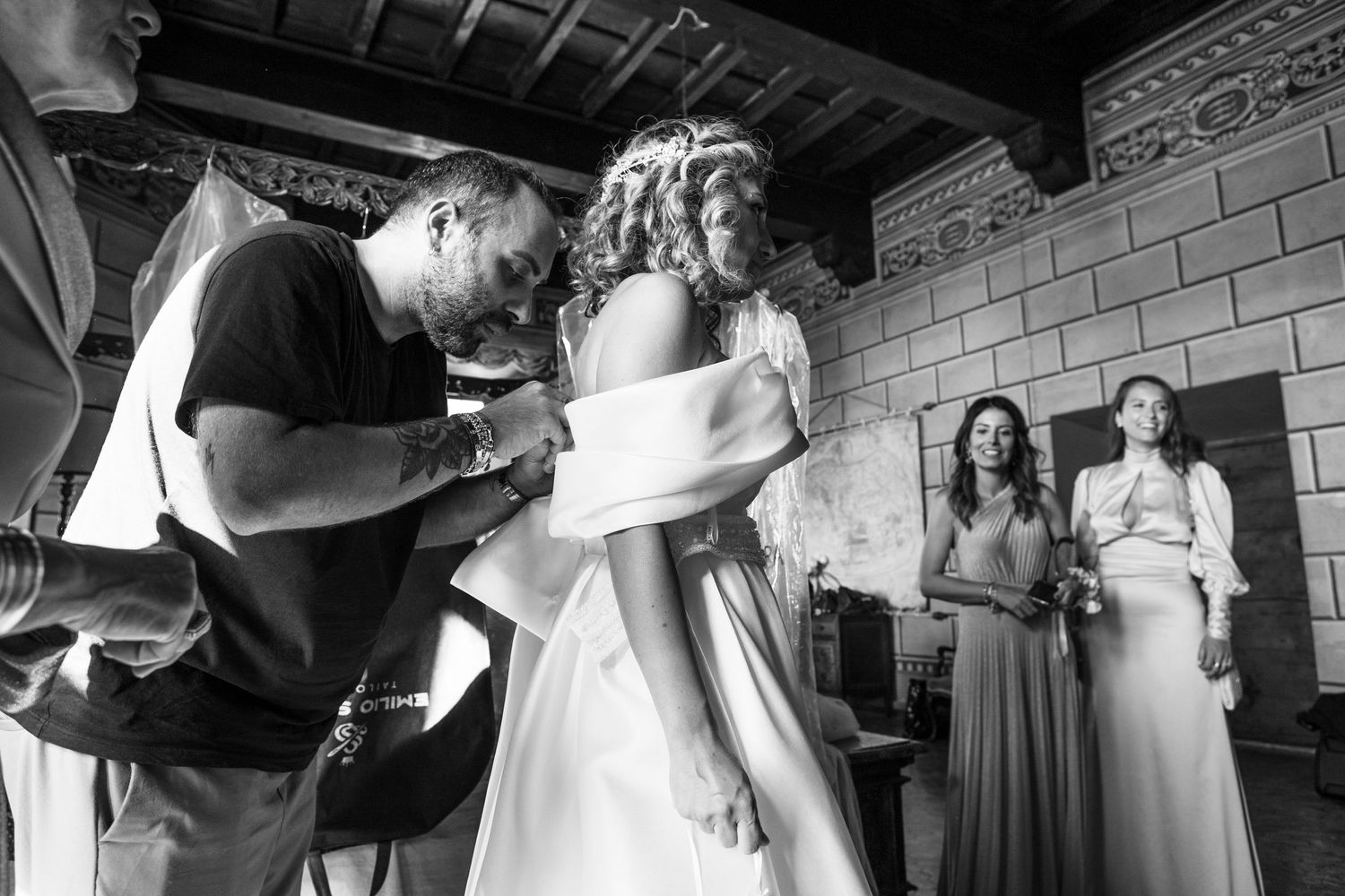 Behind-the-scenes black and white photo showing final wedding dress adjustments with bridesmaids watching.