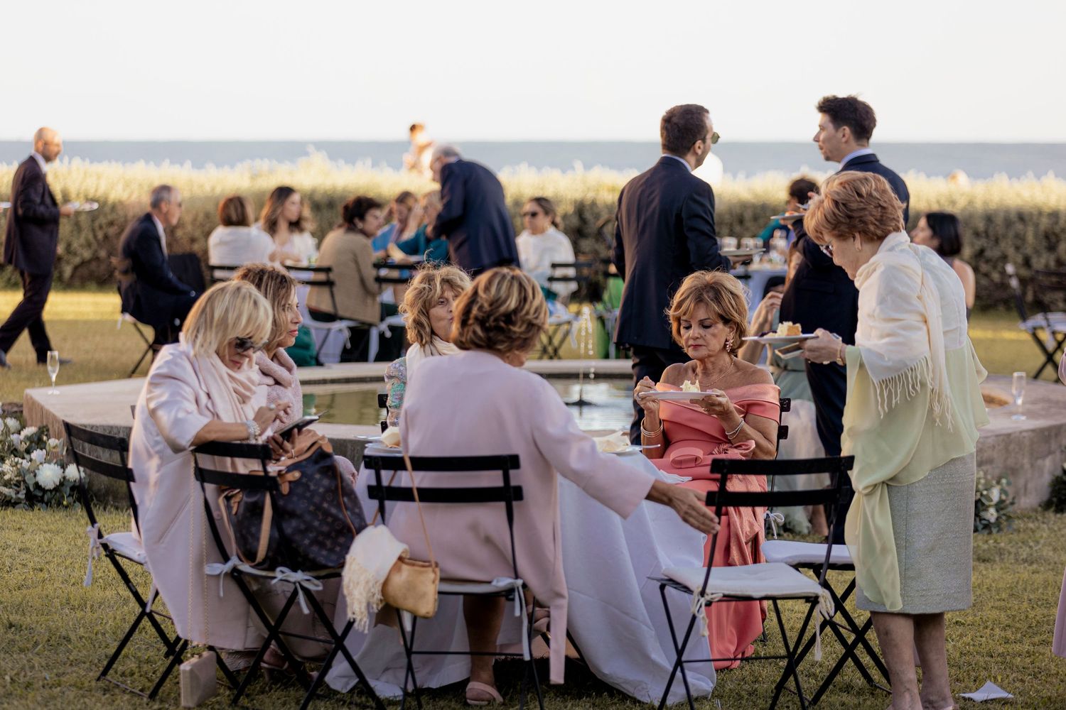 Outdoor wedding reception with guests dining at tables on a lawn at sunset.