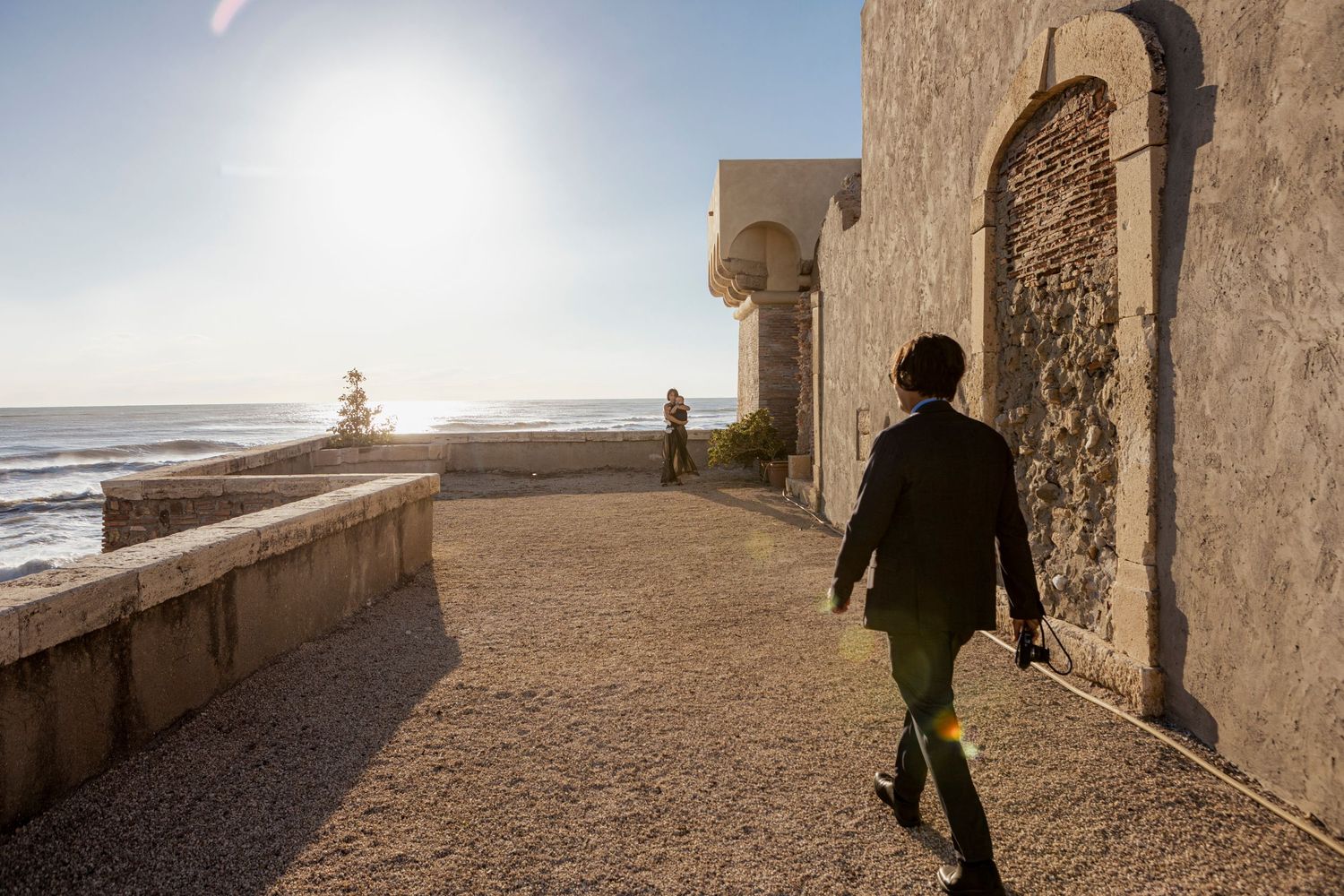 Person walking along a seaside path next to an ancient stone wall during sunset.