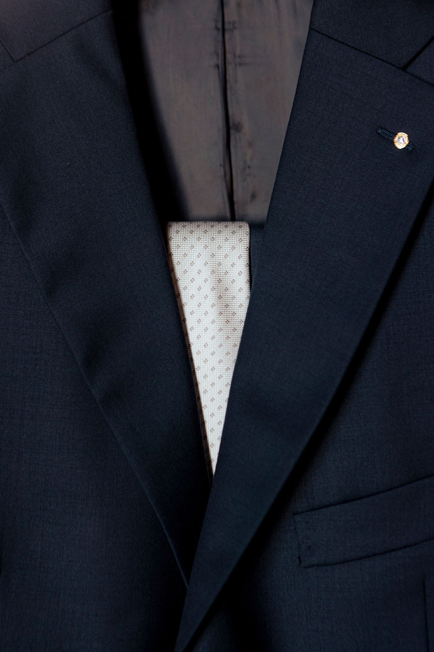 Close-up detail of a dark navy suit jacket with white patterned tie and lapel pin.