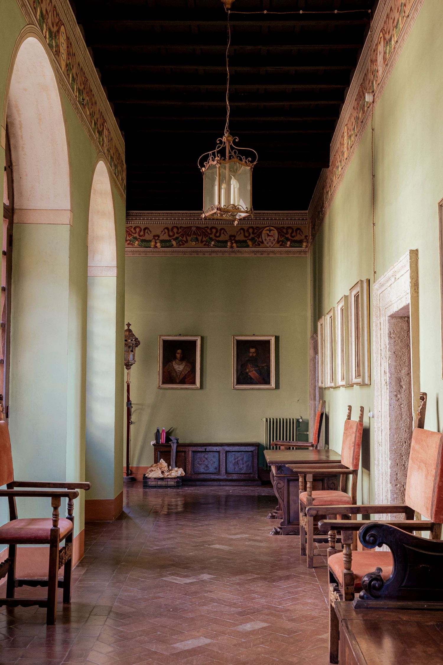 Elegant hallway with arched windows, antique furniture, and classical portraits hanging on pale green walls.
