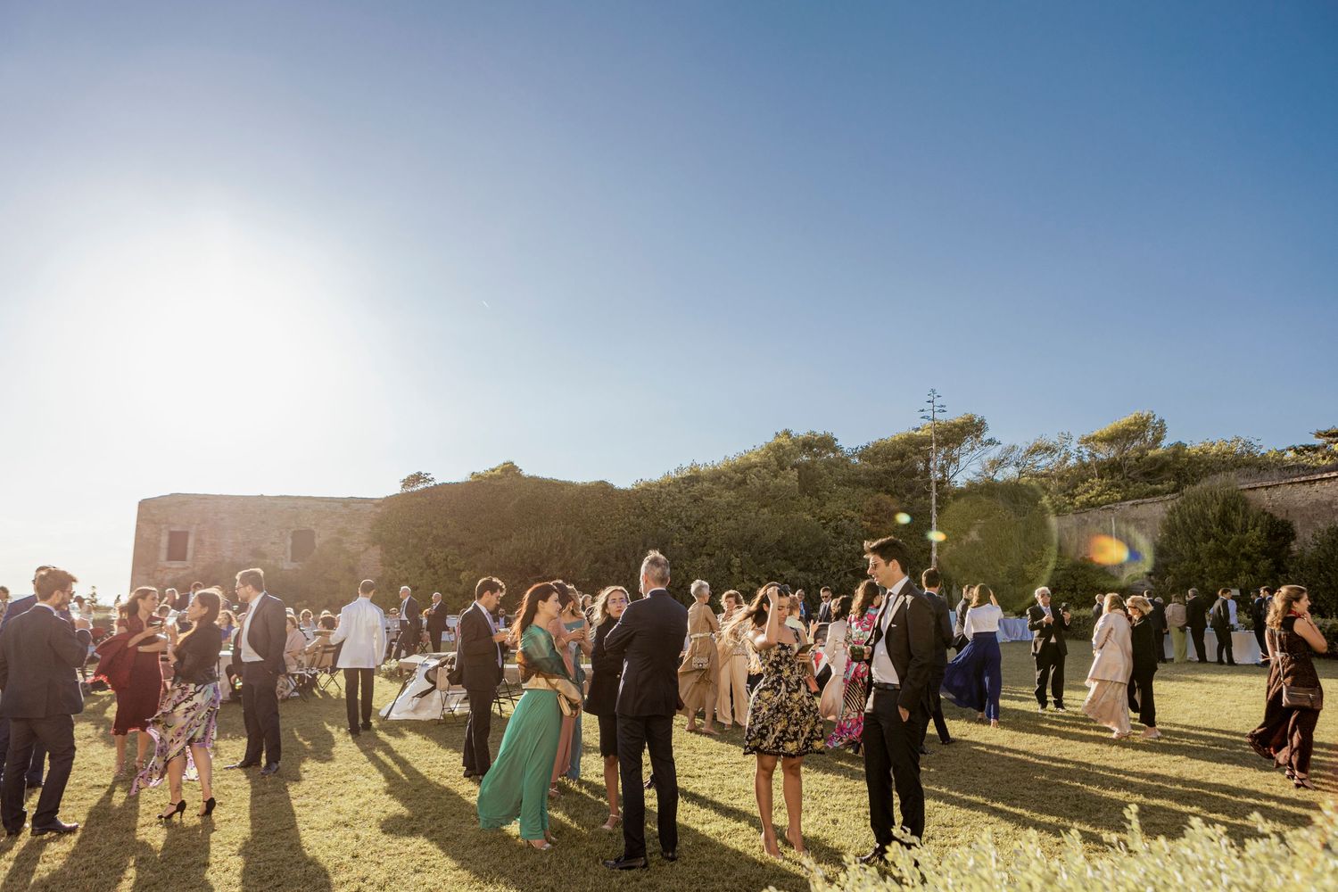 Guests gather outdoors at an elegant wedding venue during golden hour with the sun setting in the background.