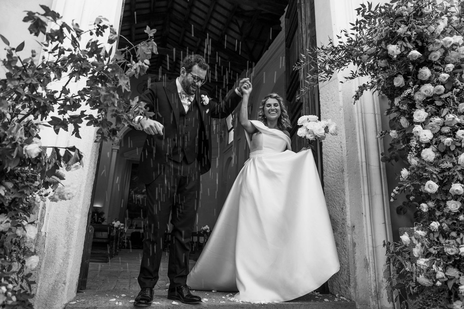 Joyful black and white moment of newlyweds celebrating with rice being thrown at church exit.