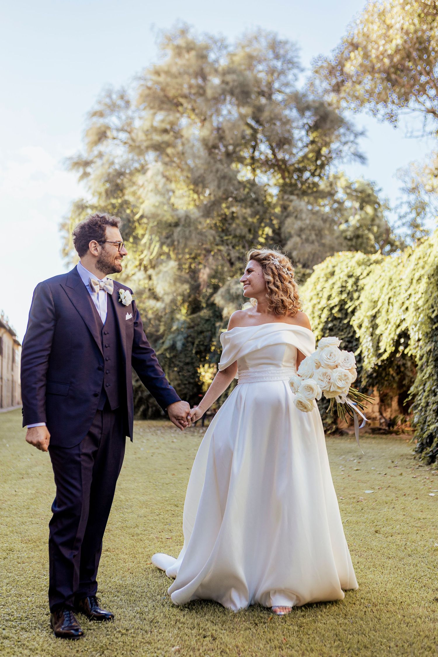 Elegant bride and groom holding hands in a sunlit garden setting.