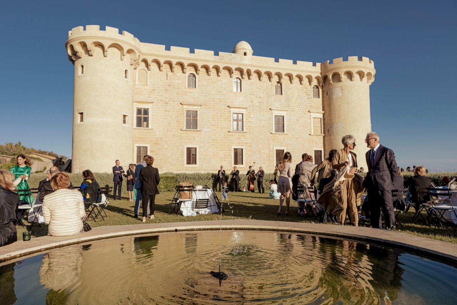Historic castle venue with stone turrets and reflecting pool where wedding guests mingle on a sunny day.