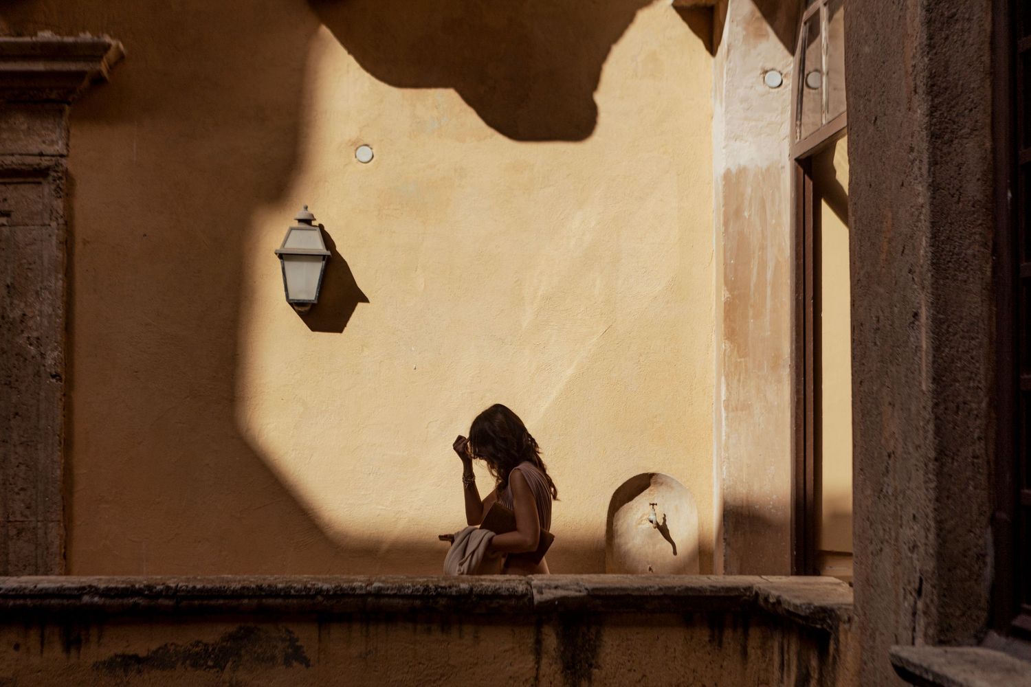 Silhouetted figure seated on a ledge against a beige wall with dramatic lighting and shadows.