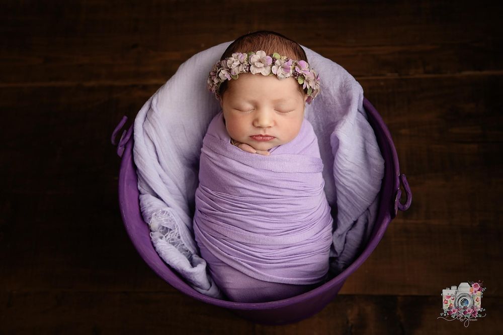 Sleeping newborn in purple wrap and delicate headband posed in round purple bowl.