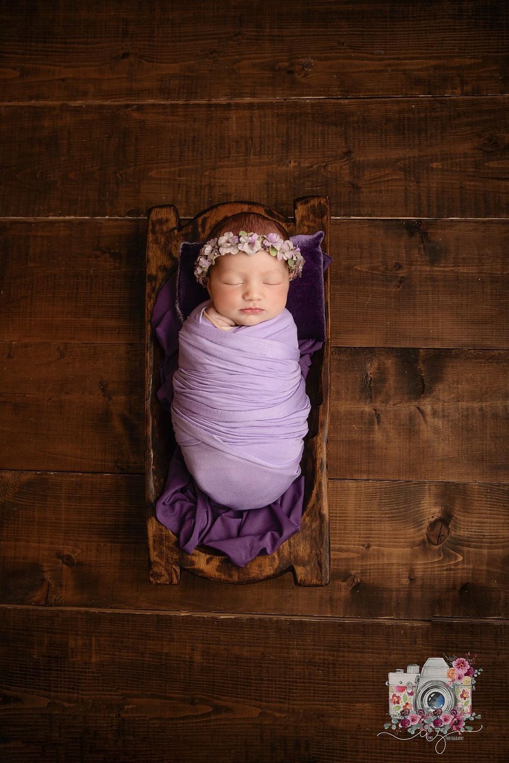 Sleeping baby wrapped in purple swaddle with floral headband posed in rustic wooden box.
